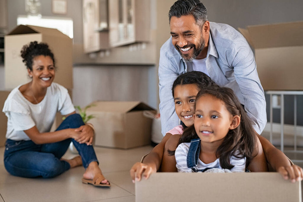 Happy children enjoying cardboard ride with indian father in new home. Cheerful daughters sitting in carton box while man pushing them. Smiling parents with two children having fun at new house in moving day. - Fotoğraf, Görsel