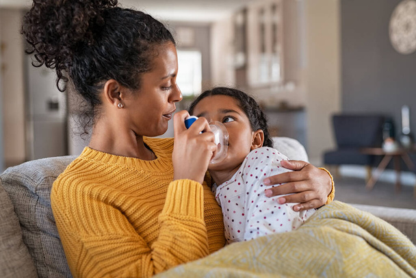 Black mother helping sick daughter use nebulizer while embracing her on couch at home. Woman makes inhalation with equipment to indian girl. Ill child lying on couch having respiratory illness helped by lovely mom. - Фото, изображение