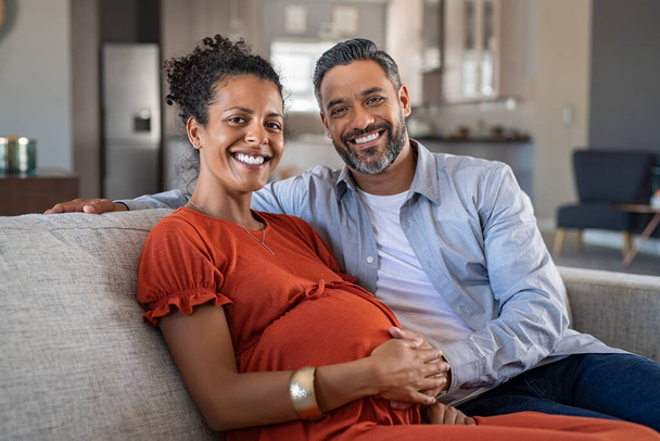 Happy indian man embracing his pregnant wife and looking at camera. Loving husband sitting with pregnant african american wife at home with hand on stomach. Portrait of excited mid adult couple expecting a baby and holding hands. - Φωτογραφία, εικόνα