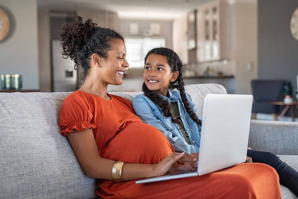 Pregnant black woman working on laptop while sitting on sofa and smiling to daughter. Cheerful expecting mother and girl relaxing on couch and using laptop to book vacation. Happy woman with baby bump and daughter working on computer from home during - Foto, Bild