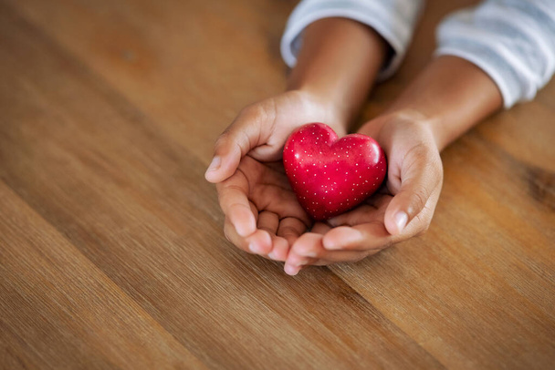 Close up of girl hands holding a red heart shape on wooden table with copy space. Top view of little cute kid hands showing small red heart. High angle view of child holding a heart-shaped stone at home: love, insurance and organ donor concept. - Photo, image