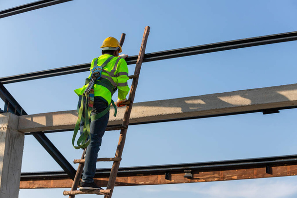 Trabajador de construcción de techo asiático usa equipo de altura de seguridad subiendo las escaleras para instalar el marco del techo, dispositivo de parada de caída para el trabajador con ganchos para el arnés del cuerpo de seguridad en el sitio de construcción. - Foto, imagen