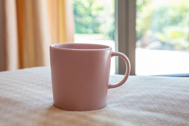 ベッドの上のコーヒーマグカップ、寝室の背景。ホットドリンクカップモックアップ。ハンドル広告付きピンク色の空白のカップ、ブランディングテンプレート - 写真・画像