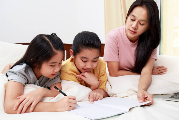 Aziatische familie moeder haar dochters doen huiswerk samen op bed in de slaapkamer. familie en thuis leren concept. - Foto, afbeelding