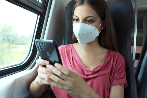 Relaxed woman with KN95 FFP2 face mask using smart phone app. Train passenger with protective mask traveling sitting in business class texting on mobile phone. Travel safely on public transport. - Photo, Image