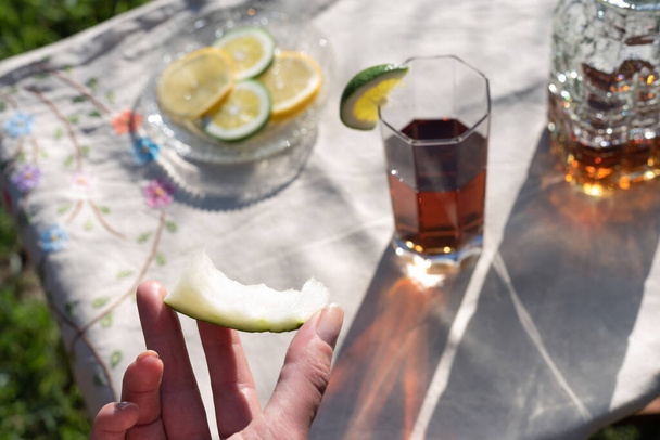 Woman hand holding eaten melon slice and ice tea sunset table background. Glass and bottle make beautiful orange shadows on linen tablecloth. Lemon and lime slices dish. Hydrate with drink and food - 写真・画像