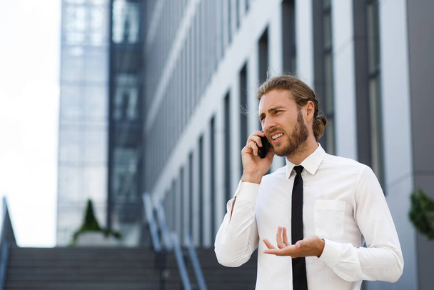 Portrait of a successful young businessman. A curly-haired man in a white shirt with a telephone against the background of a modern business center - Foto, imagen
