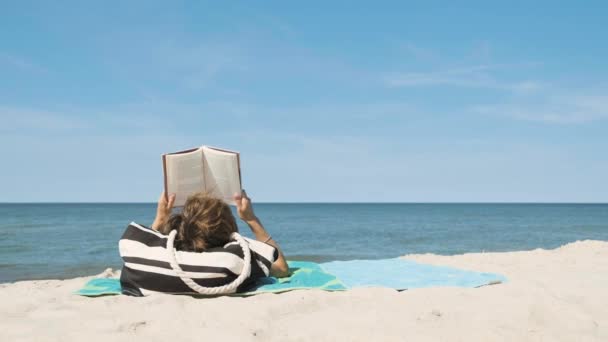 Ανώνυμη γυναίκα από πίσω κάνει ηλιοθεραπεία στην παραλία και διαβάζει ένα βιβλίο την καλοκαιρινή μέρα - Πλάνα, βίντεο