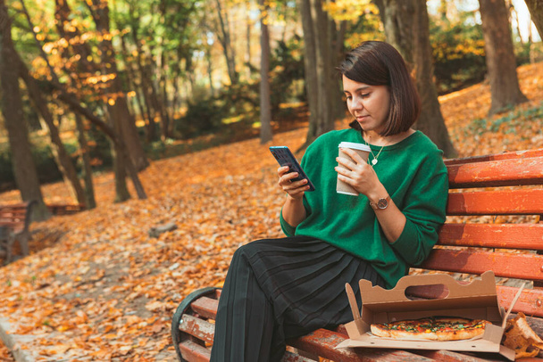молодая довольно стильная женщина сидит в осеннем парке и пьет кофе в интернете по телефону. копировальное пространство - Фото, изображение
