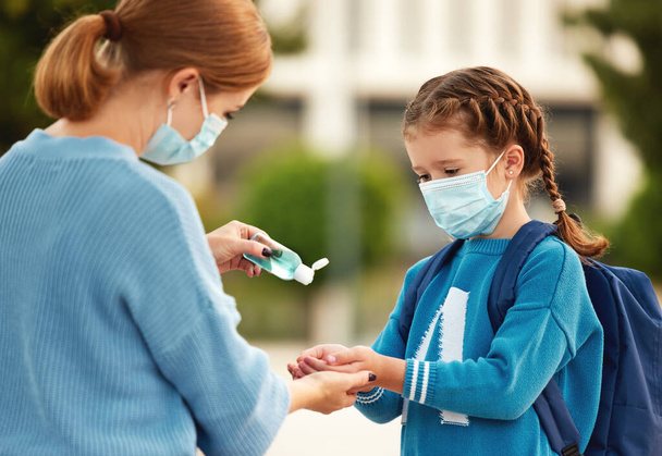 Side view of careful mom in protective mask pouring sanitizer into hands of little school aged daughter during coronavirus pandemi - Photo, Image