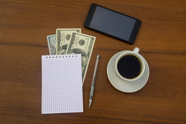 Cup of coffee, smartphone, dollars, notepad and pen on wooden desk. Financial planning concept - Photo, Image