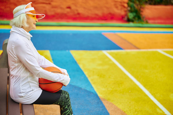 Alte, gesunde und fröhliche Dame mit Basketballball auf einem bunten Platz im Freien - Foto, Bild