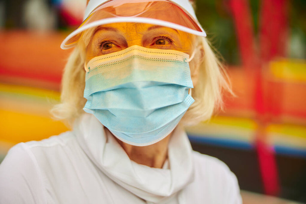 Old, healthy and cheerful lady on a colourfull square outdoors wearing mask  - Photo, Image