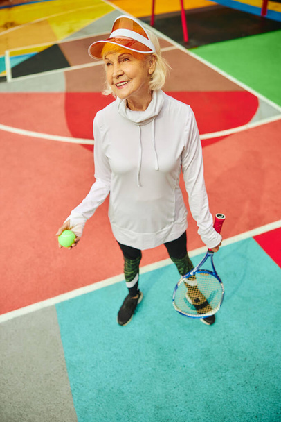 Γέρος, υγιής και χαρούμενη κυρία σε ένα χρωματιστό τετράγωνο σε εξωτερικούς χώρους και κρατώντας ρακέτα τένις με μπάλα - Φωτογραφία, εικόνα