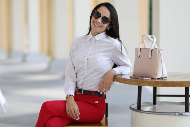 Стильна жінка сидить в сонцезахисних окулярах, посміхаючись біля столу лежить смартфон і стоїть сумка
 - Фото, зображення