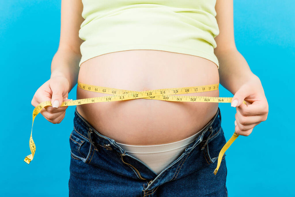 Primer plano de una mujer embarazada en jeans abiertos midiendo su vientre con una cinta métrica en un fondo colorido con espacio para copiar. Medición de pulgadas. - Foto, Imagen