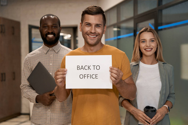 Fiatal boldog kaukázusi férfi, irodai dolgozó, papírt mutat szöveggel VISSZA AZ AJÁNLATBA a kamera előtt és mosolyog, miközben kollégáival áll az irodában - Fotó, kép