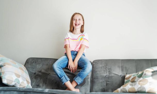 A cute little girl 8 years old in a striped T-shirt and jeans sits on a gray sofa at home and smiles happily - Φωτογραφία, εικόνα