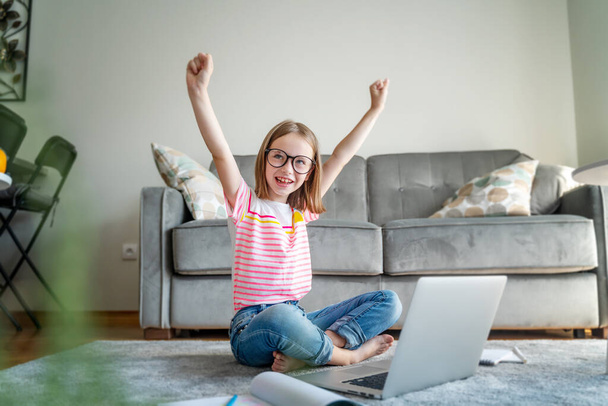 Happy cute little girl 8 years old in a striped t-shirt and jeans with glasses sits at home on a carpet in front of a laptop, remote education technologies and homework - Foto, Bild