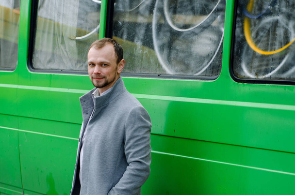 Shot of a Handsome businessman freelancer in grey coat and biege trousers smiles and standing near green bus - Фото, зображення