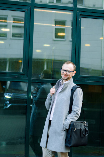 Handsome businessman freelancer in grey coat and biege trousers smiles and walking down the sidewalk - Fotoğraf, Görsel