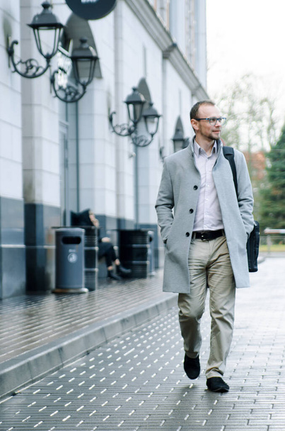 Handsome businessman freelancer in grey coat and biege trousers smiles and walking down the sidewalk - Foto, afbeelding