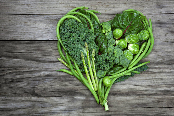 Le verdure verdi biologiche sane si uniscono per fare un concetto sano del cuore. Le verdure includono cavolo, fagiolini, asparagi, cavoletti di Bruxelles e varie lattughe su uno sfondo rustico di legno. - Foto, immagini