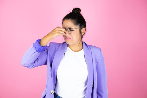 Young beautiful business woman over isolated pink background smelling something stinky and disgusting, intolerable smell, holding breath with fingers on nose - Photo, Image