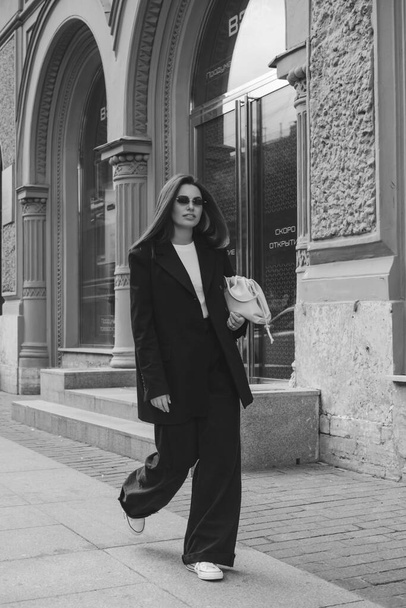 Outdoor fashion portrait of a  young woman in a trouser suit. Streetstyle. A brunette on the streets of the city in a fashionable oversized jacket and sunglasses from the sun. - Φωτογραφία, εικόνα
