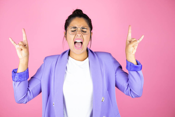 Молодая красивая деловая женщина на изолированном розовом фоне кричит с сумасшедшим выражением, делая рок-символ с поднятыми руками - Фото, изображение