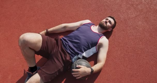 Giocatore di basket che riposa dopo la partita, sdraiato con la palla sul pavimento del campo all'aperto - Filmati, video