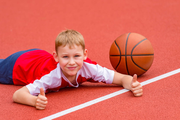 Chlapec pózuje s basketbalovým míčem na umělém červeném trávníku. Sportovní trénink v terénu. Kid sport online koncept - Fotografie, Obrázek