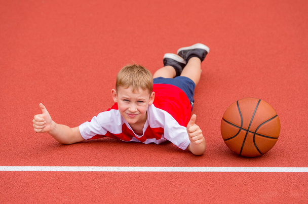 Chlapec pózuje s basketbalovým míčem na umělém červeném trávníku. Sportovní trénink v terénu. Kid sport online koncept - Fotografie, Obrázek