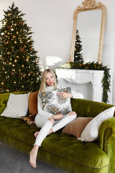 A beautiful model girl sits on a couch near the Christmas tree in the decorated for New year living room interior - Φωτογραφία, εικόνα