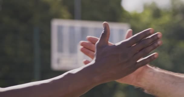 Καυκάσιοι και Αφρικανοί άνδρες που κρατιούνται χέρι-χέρι ως σύμβολο ενότητας στο υπαίθριο γήπεδο μπάσκετ - Πλάνα, βίντεο