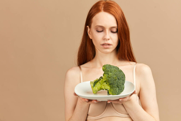 Anorexic woman in underwear holding plate with raw broccoli isolated in studio - Foto, Bild
