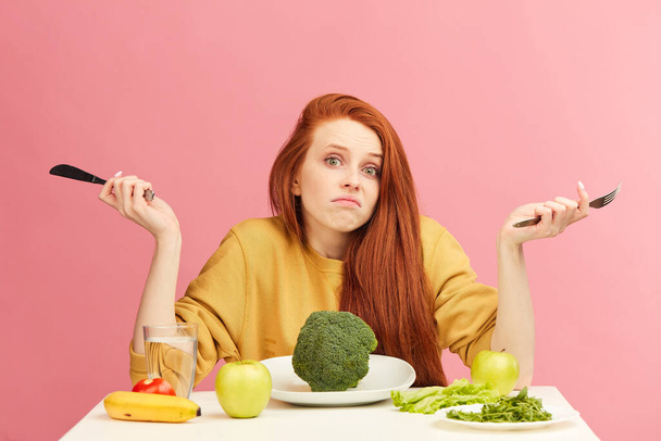 Puzzled, uncertain young red-haired woman sitting at table, served with broccoli - Fotó, kép