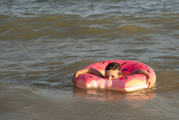 Een 5-jarige jongen zwemt in de zee, verheugt zich en heeft plezier in een opblaasbare ring in de vorm van een donut - Foto, afbeelding