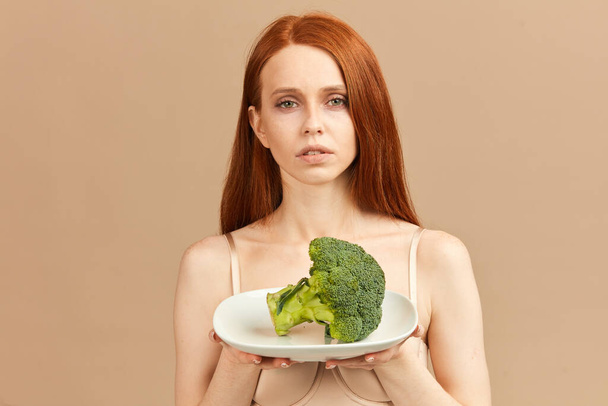 Anorexic woman in underwear holding plate with raw broccoli isolated in studio - Photo, image