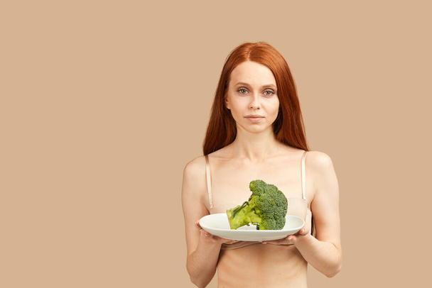 Anorexic woman in underwear holding plate with raw broccoli isolated in studio - Фото, зображення