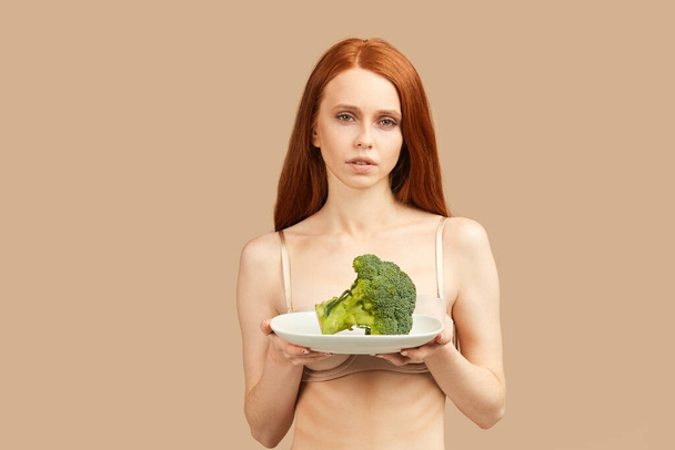 Anorexic woman in underwear holding plate with raw broccoli isolated in studio - Fotó, kép
