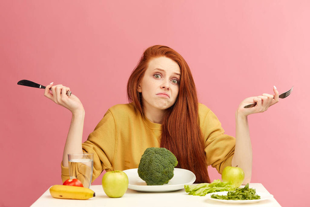 Mujer joven pelirroja, desconcertada e incierta, sentada a la mesa, servida con brócoli - Foto, Imagen