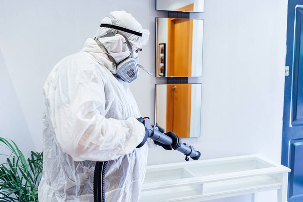 Evde PPE giyen bir adam COVID-19 'dan cam bir masayı dezenfekte ediyor. Salgın sağlık konsepti - Fotoğraf, Görsel