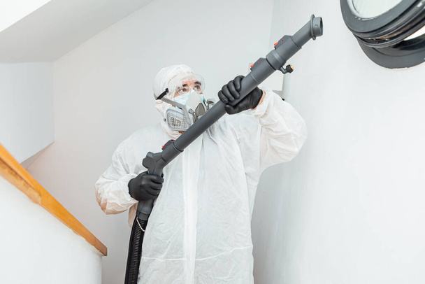 Man wearing PPE disinfecting a house next to a round COVID-19 window. Pandemic healthcare concept - Photo, Image