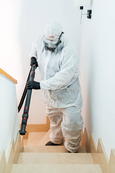 Чоловік, одягнений в PPE, піднімається по сходах будинку, дезінфікується від COVID-19. Концепція пандемії охорони здоров'я
 - Фото, зображення