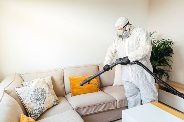 Чоловік у PPE дезінфікує вітальню будинку з дезінфікуючим апаратом COVID-19. Концепція пандемії охорони здоров'я
 - Фото, зображення
