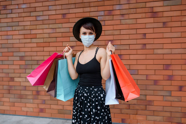 Junge Frau mit Schutzmaske hält nach einem guten Einkaufstag viele bunte Einkaufstüten in der Hand. Einkaufen in der Coronavirus-Covid-19-Pandemie.  - Foto, Bild
