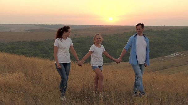healthy smiling family holding hands walking in field at sunset in the mountains, daughter holding mom and dad by hands. happy child and parents walk in rays of beautiful sun, travel on vacation - Photo, Image