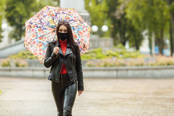 Una chica enmascarada camina por la calle. Una chica con una máscara protectora camina por el parque con un paraguas bajo la lluvia. Infección por Coronavirus COVID-19. - Foto, Imagen