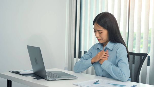Młoda Azjatka siedzi w pracy i odczuwa ból w klatce piersiowej spowodowany stresem. - Zdjęcie, obraz